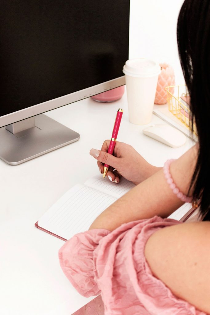 woman in a pink dress writing a note in front of desktop computer