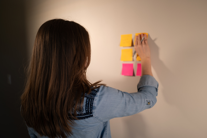 Chynna is facing the wall, we can see her backside. She is wearing a blue long sleeve shirt, her hair is straight down and she is placing sticky notes on the wall to help with her planning.