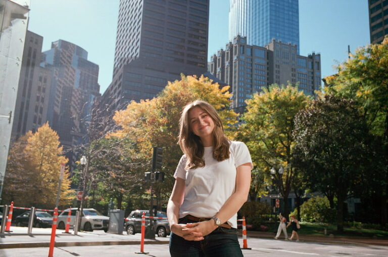 Chynna standing in the street wearing a white t-shirt. Her hair is down and she is slightly looking down at the camera, hands folded into one another and smiling