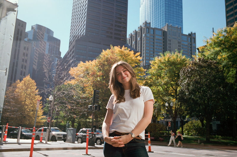 Chynna standing in the street wearing a white t-shirt. Her hair is down and she is slightly looking down at the camera, hands folded into one another and smiling