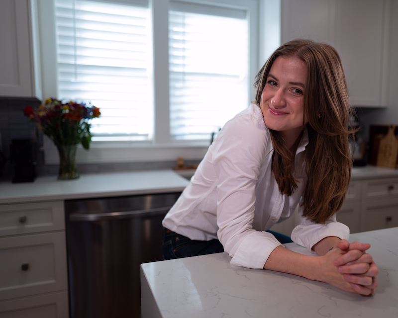 Chynna Benton in her kitchen wearing a white button up shirt and her brown hair worn down. She is bending onto her kitchen island with her hands into on another smiling at the camera.