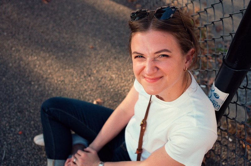Chynna sitting on the ground looking up at the camera. Sitting on the floor crossed legged, hair up in a clip, black sunglasses on her head and she is scrunching her face to the camera.