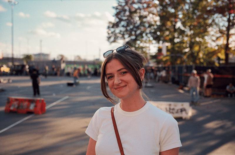 Chynna standing in a public park with her hair up in a clip, black sunglasses on the top of her head with two pieces of hair in front of her face. She is wearing a white t-shirt with a brown purse crossed over her body.