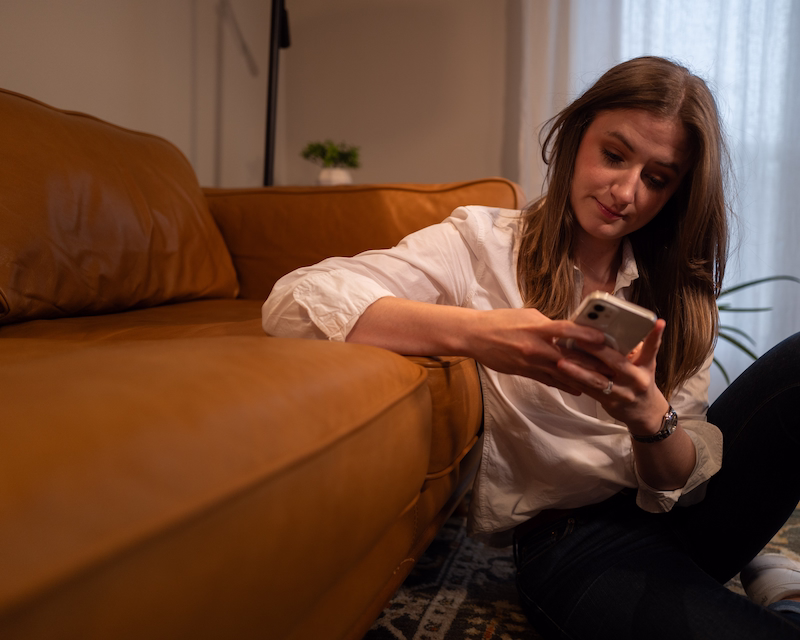 Chynna Benton sitting on her living room floor. Leaning on her brown couch. Wearing a white button up shirt and her hair straight down. As she is sitting on the floor she is looking down at her phone doing work.