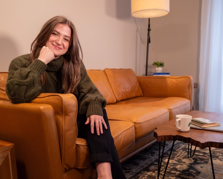 Chynna Benton in a green sweater, sitting at the end of her brown couch in her licing room. Her head is resting onto her fist that is propped onto the end of couch and she is softly smiling right at the camera.