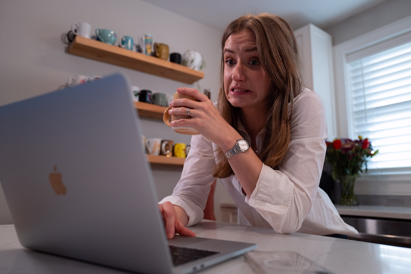 Chynna Benton leaning onto her kitchen island, looking at her computer with a worried face. She is holding a coffee cup in her left hand, scrolling on the computer.