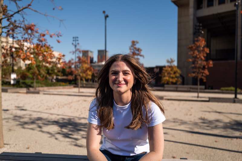 Chynna Benton sitting on a bench in a park. Wearing a white tshirt, her hair is down, and she is slightly smiling to the camera.