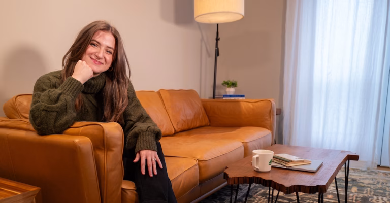 Chynna Benton sitting on her brown couch. Wearing a long green sweater resting her head on her hand as she smiles to the camera