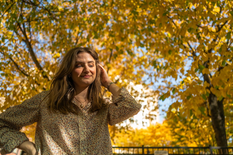 Chynna wearing a polkadot shirt and pushing her hair back in front of yellow fall trees