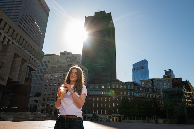 Chynna Benton drinking an iced coffee in a city square with the sun shining in the background